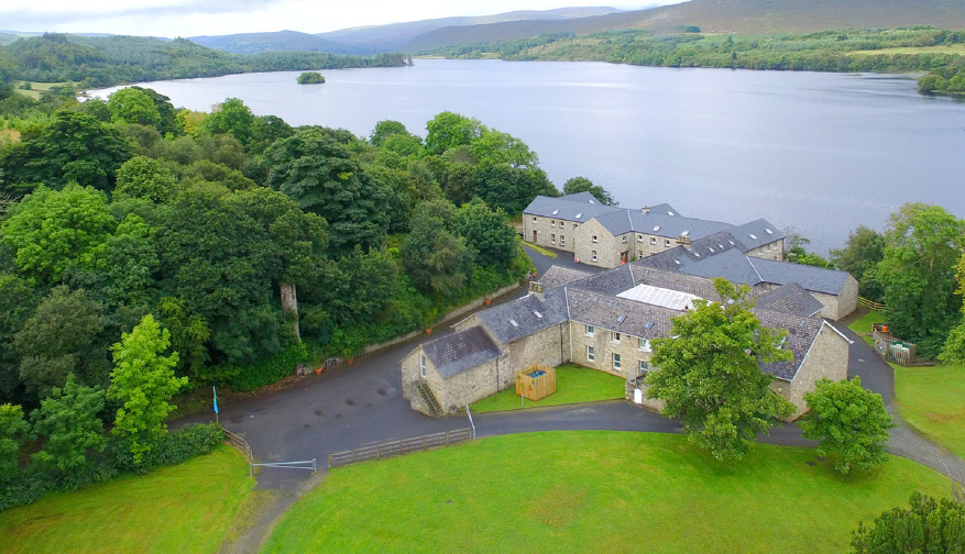 Oblique aerial image of a large, L-shaped building in front of a lake, among forestry