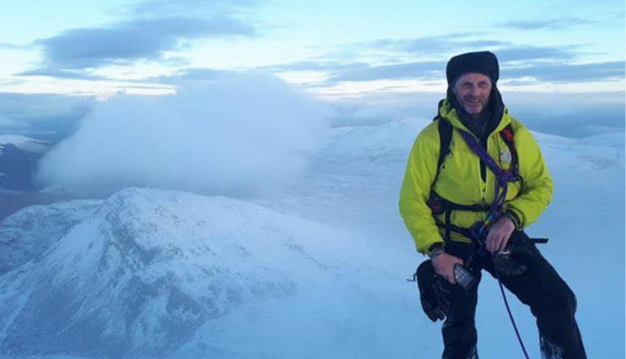 Man atop a snowy mountain peek among clouds. He is wearing a luminous yellow jacket and a black hat and has a smartphone in his hand