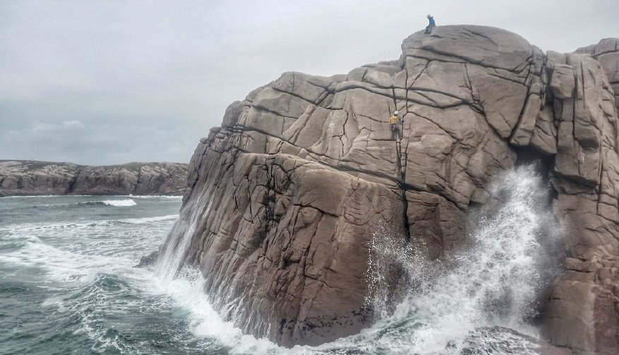 Two climbers scaling along a cliff, with waves crashing down below